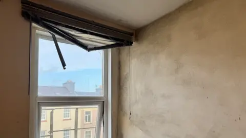 There is a window to the left with a blind rolled to the top but broken with slats hanging down. The corner of the room with a large cream wall to the left is dappled with patch black mould. You can see townhouses through the window outside and a blue sky and sea in the distance.