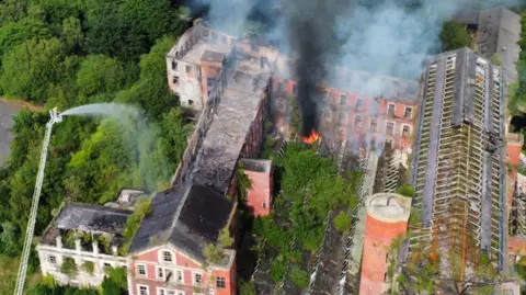 NIFRS Another aerial photograph of the HIlden Mill fire in June 2021. The image shows smoke rising from derelict Victorian buildings and firefighters using an aerial hose to tackle the flames. 