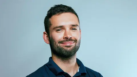 RNLI Ted Morgan smiles at the camera. He has short dark hair and a dark beard. He wears a navy polo t-shirt and stands against a light blue background. 