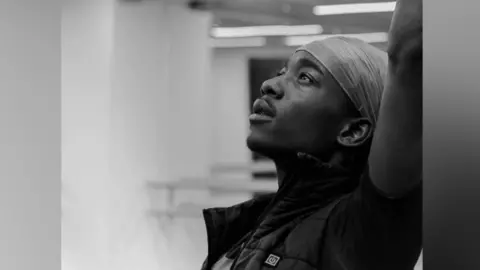 Nahum McLean/Jack Thomson Adult Nahum stands in a dance studio looking up to the ceiling with one hand stretched into the air.