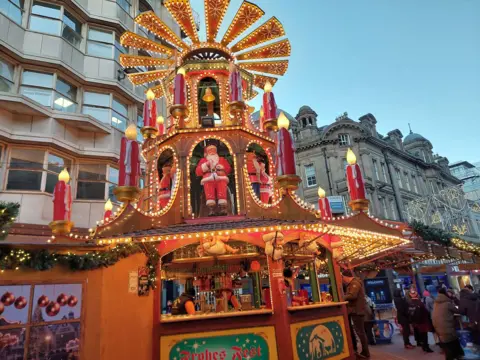 A stall selling alcohol at a Christmas market. The wooden stall is adorned with golden fairylights, giant fake red candles, a bell, Santa decorations