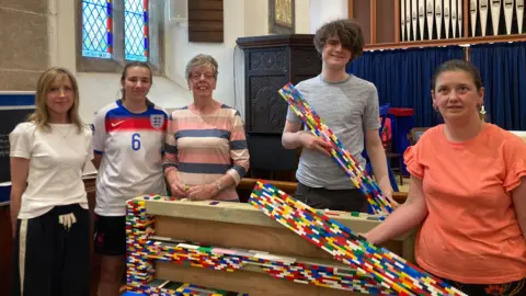Five people working on a life sized Lego bench. Working left to right, lady in white top with black trousers, student in a sports kit, lady with pink blue and white striped top, boy with grey t shirt holding part of the Lego structure and lady in orange top.