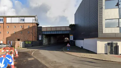 Google A road underneath a railway bridge. There are buildings on either side of the bridge and roadworks on the far left. 