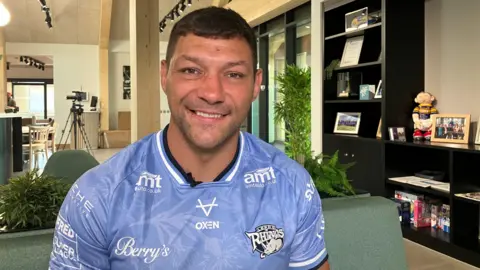 Ryan Hall, who has short dark hair and dark eyes. He is smiling in the photo and wearing a blue Leeds Rhinos jersey.