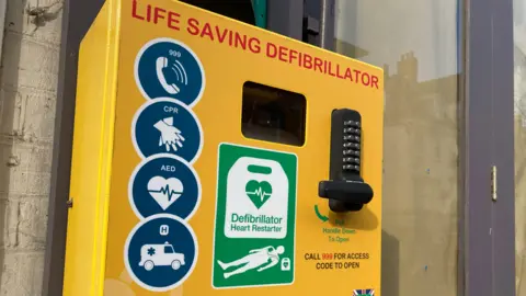 Andy Trigg/BBC A defibrillator in a yellow box attached to a wall