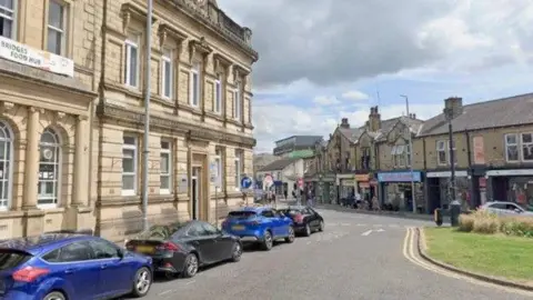 Four cars parked along a road in Brighouse, opposite a parade of shops.
