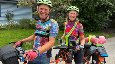 Chris and Nicky - dressed in multi-coloured cycling T-shirts and neon green helmets - are sitting on their orange tandem, which has luggage on the back. They are on a country road with greenery and a pale blue-painted house, partially visible, in the background