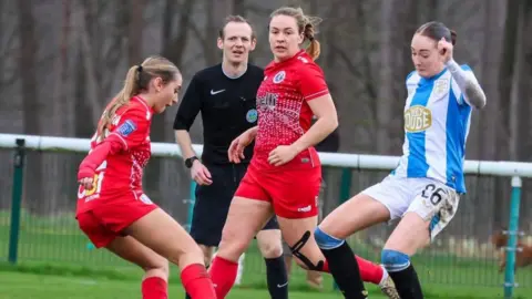 supplied A women's football match. Two girls in red compete to win the ball from an opposition player in blue and white. A referee in black watches closely