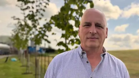 John McClenaghan pictured on a farm. He is bald and wearing a purple lined shirt with brown buttons. He is looking directly into the camera. 