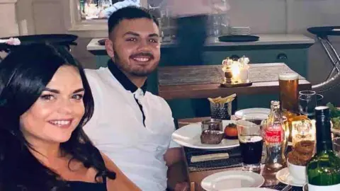 Aime Hewitt Kye and Aime smiling while sitting in a restaurant. Aime has long, dark brown hair. Kye has dark hair and a dark beard. He is wearing a white polo shirt with buttoned-up dark collar.