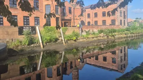 Water from the canal looks low at the sides due to mud and the wooden sticks being visible where the water would have normally been. There is a building behind the canal with orange bricks and black windows. The picture is taken on a hot day with a clear blue sky that reflects on the water. 