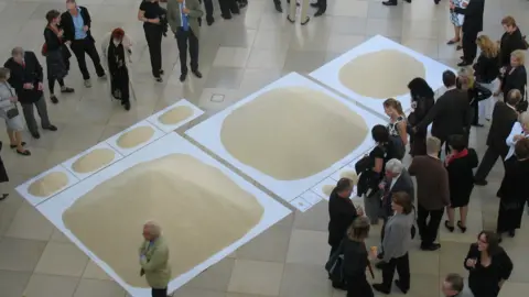 There are three large white squares placed diagonally across the floor, each with different amounts of rice piled on top of them. The first on the left has the most rice piled up, and it goes down in descending order. Above the first square are four rectangles in a line next to each other with similar amounts of rice on them. There are people gathered around either side of the displays, some talking to one another and others looking on at the piles