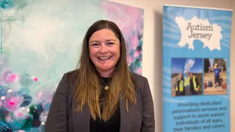 BBC Emilie smiles at the camera standing next to one of her paintings and a Autism Jersey banner