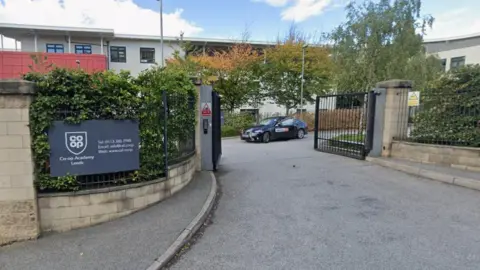 The entrance to a school, as marked by an open set of black gates. A sign with the school's name is on the fence to the left of the gates. A taxi is emerging from the other side.