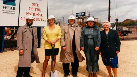 Handout by Roger Clubley Roger Clubley, left, stands next to those who helped make the National Glass Centre a reality in a photograph taken around 1996. They are standing on land which has been redeveloped near the River Wear and now features beige coloured stones, and one person holding a black handled silver spade. Clubley is standing in front of a large white sign that reads 'Site of the National Glass Centre' and is held into the ground with large blue poles.