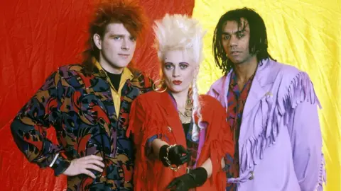 Getty Images The band poses for a publicity photograph against a backdrop divided into red and yellow sections. Tom Bailey wears a patterned jacket in red, purple and black tones, Alannah Currie wears a bright red outfit with large gold jewellery and black gloves, and Joe Leeway wears a light purple fringed jacket over a patterned shirt. The group are backstage at the Pine Knob Music Theatre in Clarkston, Michigan, on 16 August 1987.