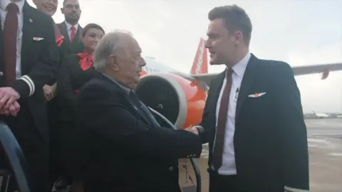 Ant Saddington/BBC An older man is shaking hand with a younger man on the steps of a plane. Both are smartly dressed.
