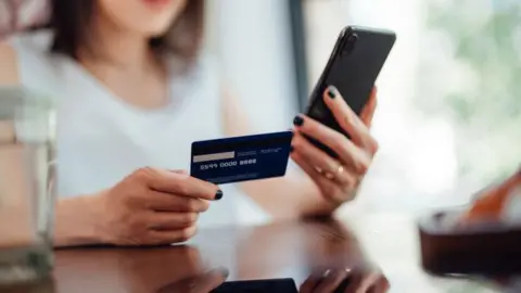 A woman is holding a credit card or debit card and doing online shopping on a smartphone. 