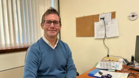 Andy Abrahams is pictured wearing a blue jumper with a white shirt underneath. He has dark hair and wears glasses. He is sat at a desk.