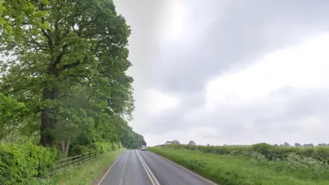 Google A rural North Yorkshire road. On the left are trees and a fence dividing the grass verge from private land. On the right are fields, separated from the grass verge by a hedgerow.