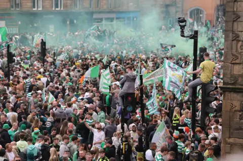 PA Media A large crowd of Celtic supporters gathers in a Glasgow street, with green smoke in the air and flags being waved, while some people sit on traffic lights.