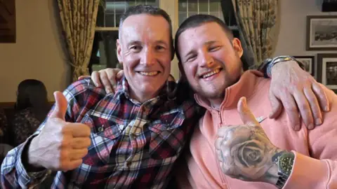 A father and son with one arm around one another and the other doing a thumbs up sign to the camera 