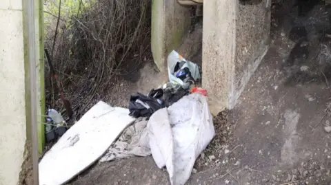 Crown Prosecution Service A makeshift camp under a small bridge in Brantham. A white crumpled up duvet can be seen alongside a bag of rubbish and other items stress across the ground 
