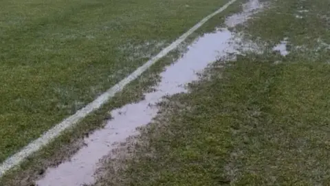 A close up of a long puddle in waterlogged ground on a grassy football pitch. The water runs parallel to a painted white line on the pitch.