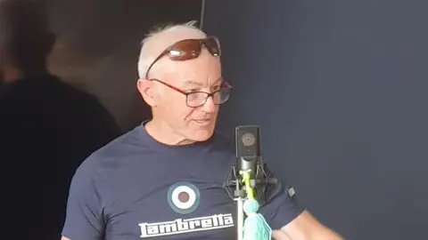 Andrew Mears is standing in front of a microphone and speaking animatedly. He has short grey hair and is wearing black glasses. He also has a pair of sunglasses resting on top of his head.