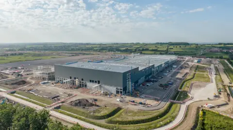 A large grey factory building in construction on a building site surrounded by trees and green fields on a sunny day