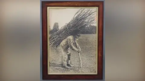 Denhams A photo of a man hunched over in a field with sticks strapped to his back.