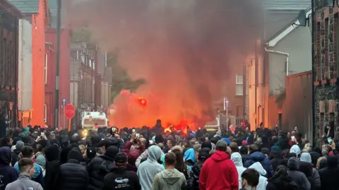 A crowd of hundreds or people in a street looking towards police vehicles and a part of the street that is one fire