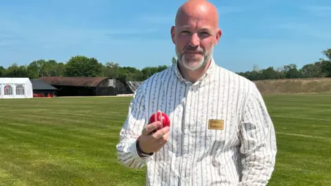 Kayne is standing on a cricket pitch holding a cricket ball in his right hand. He is wearing a white shirt with thin lines down it. He has a short white and grey beard.