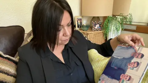 A woman in a black jacket and shirt sits on a sofa looking at a photo of three young men.