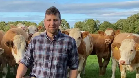 Fergal Watson in a field on his farm, surrounded by cattle. He has short, fair hair and is wearing a blue and navy checked shirt with the sleeves rolled up. There are trees in the background.