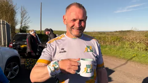 Gary McKee holding a a mug and smiling brightly at the camera. He has short grey hair and a close-cropped beard, is wearing running grey, yellow and green gear. He is standing on a rural road with a hedge and fields on the right. There are people talking behind him.