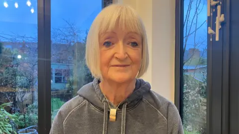 Jo James has a short blonde bob and is wearing a grey hoodie and standing in front of windows looking out onto a garden. 