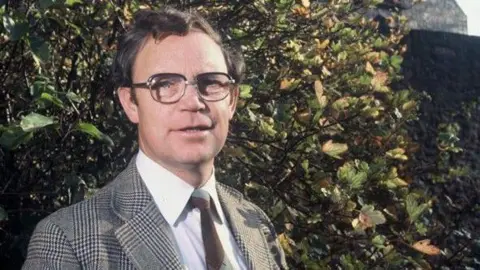 Tern TV JIm McColl in a checked tweed jacket, white shirt and brown tie, standing in front of a tree. He is smiling and looking directly into the camera