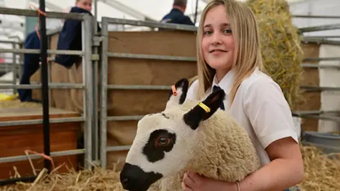 A girl with shoulder length blonde hair wearing a white short-sleeved school blouse holds a lamb in her arms. She is sitting in a pen with straw on the floor.