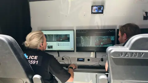 Shariqua Ahmed/BBC Two officers - a woman and a man, are watching three screens in front of them, inside the police van, with their backs to the camera.