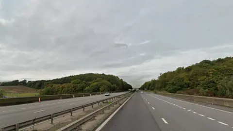  A Google streetview of both carriages of a motorway. There are cars in the distance and woodland flanking either side of the road. 