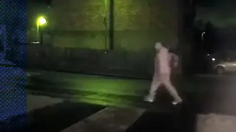 CCTV footage of a person walking across a dark street at night. There is a light at the left that provides a greenish hue to the images. The person is in grey.