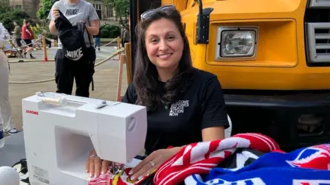 A woman with long black hair sits at a sewing machine with a pile of football scarves nearby. She smiles at the camera.