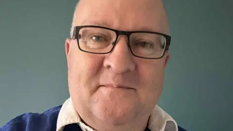 Andy Paterson A man with a bald head and glasses in a blue retro Queen of the South top with a white collar