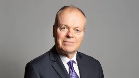 UK Parliament Clive Betts looks into the camera. He is wearing a navy blue suit, a white shirt and purple tie.