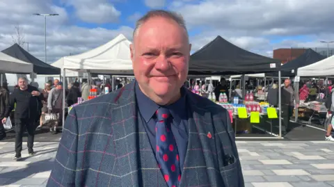 A man wearing a mainly blue jacket and waistcoat, a blue shirt and a blue tie with red circles is looking at the camera, with stalls behind him.