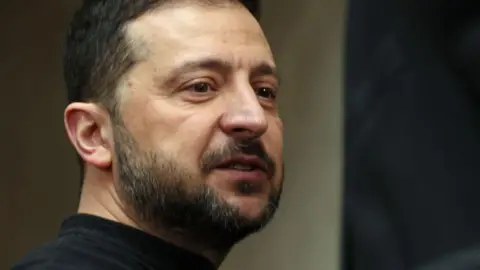 Getty Images A close-up shot of Volodymyr Zelensky, who has short brown hair, brown eyes and facial hair, and wears a black top.