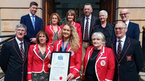 Hannah Scott is pictured with members of Bann Rowing Club. They are standing outside Coleraine Town Hall. She is holding her framed freedom of the borough certificate. 