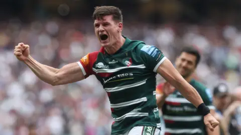 Freddie Burns holds his right arm out with his fist clenched in celebration after Leicester win the Premiership title in 2021. He is standing on the pitch at Twickenham wearing green and white striped Leicester kit