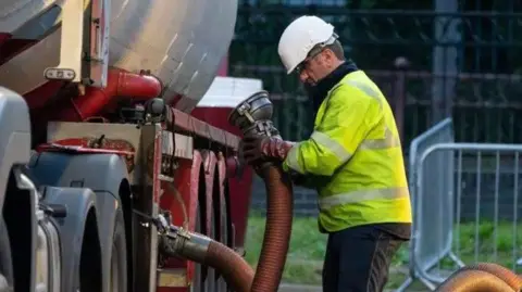 A man in a yellow high visibility jacket and white hard hat holding a fuel pipe next to a tanker lorry.